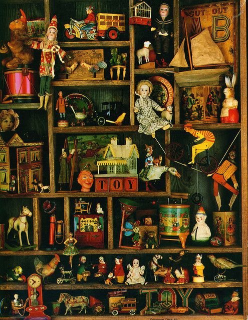 A cabinet of antique toys on wooden shelves: tin cars, dolls, a clown, a rocking horse and alphabet blocks